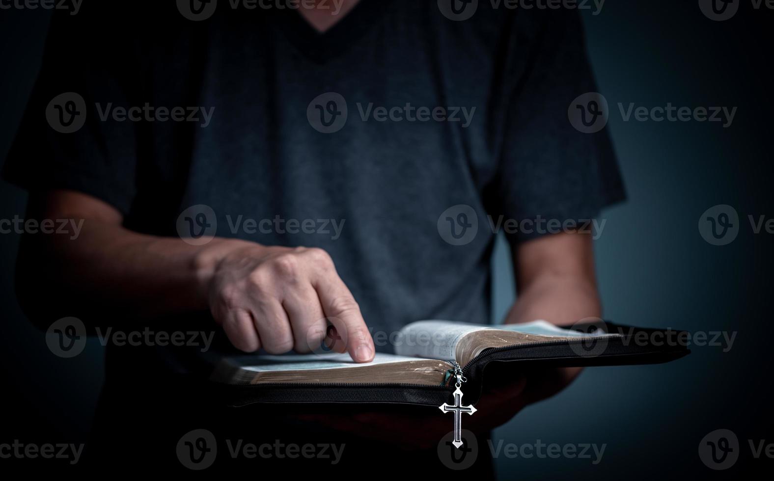 Young man reading Holy Bible and pointing on the word with cross necklace in font of the book. photo