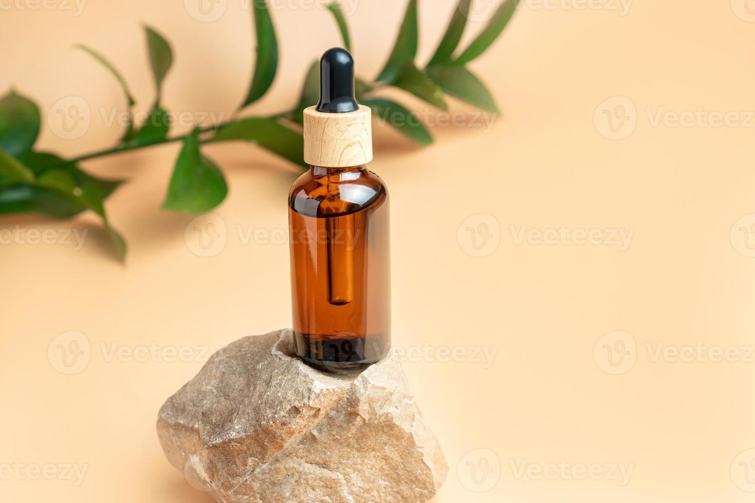 medicina natural o aceite aromático o esencia de belleza con cuentagotas en un podio de piedra con rama de ruskus verde sobre fondo beige. concepto de cuidado de suero de spa facial y corporal foto