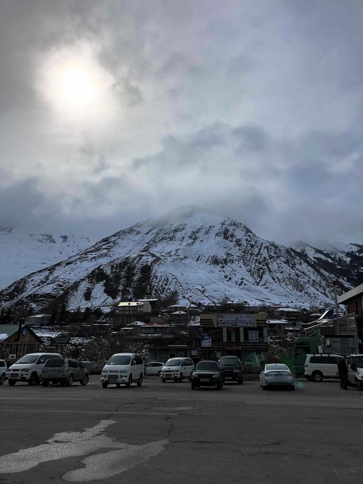 Parking and stone houses, buildings, shops, refueling on a beautiful mountain winter resort with high mountain peaks and rocks covered with snow against a blue sky. Georgia, Tbilisi, April 17, 2019 photo