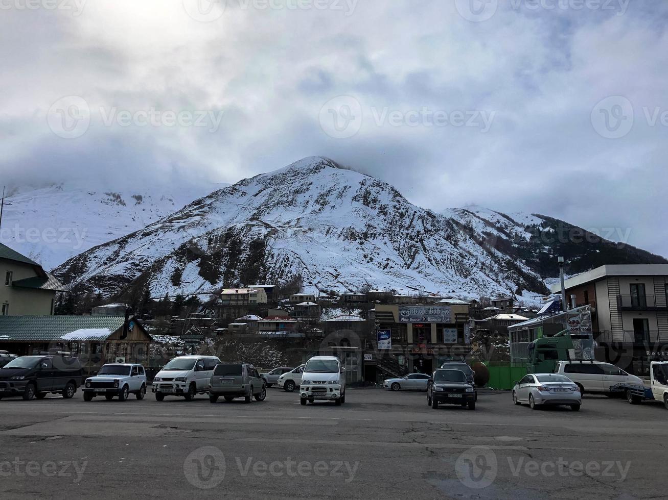 estacionamiento y casas de piedra, edificios, tiendas, reabastecimiento de combustible en un hermoso complejo invernal de montaña con picos de alta montaña y rocas cubiertas de nieve contra un cielo azul. Georgia, Tiflis, 17 de abril de 2019 foto