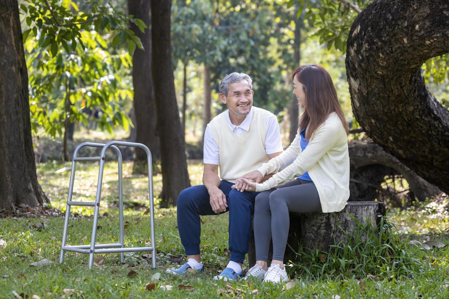 Asian senior father with walker and daughter sitting together under the big tree in the park during summer for light exercise and physical therapy concept photo