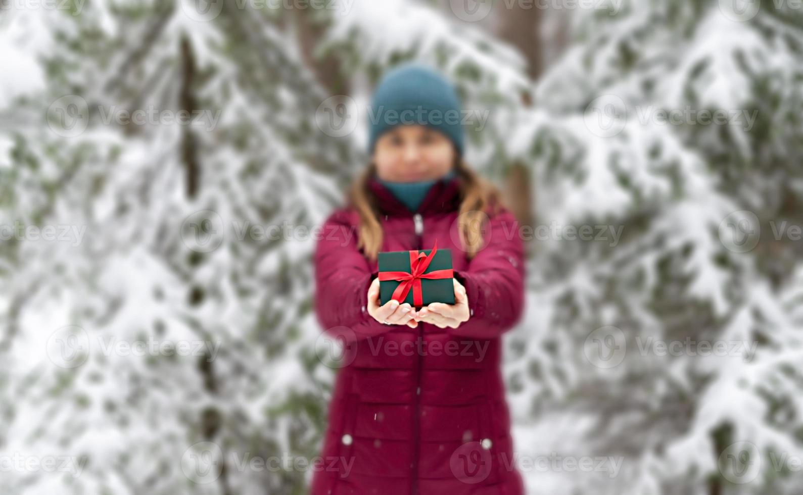 young woman in defocus in red jacket holding in hands Gift green box with red ribbon against snowy spruce tree in winter forest Christmas and New Year holidays present photo