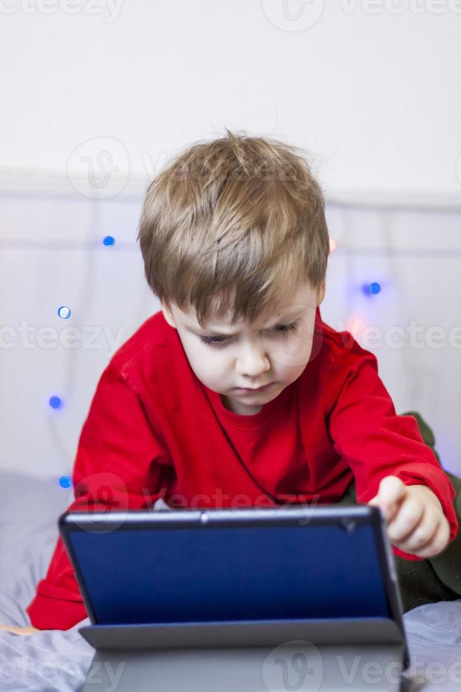 A cute boy is on the bed looking at a tablet. Online training, communication on the Internet. Funny smiling child. photo