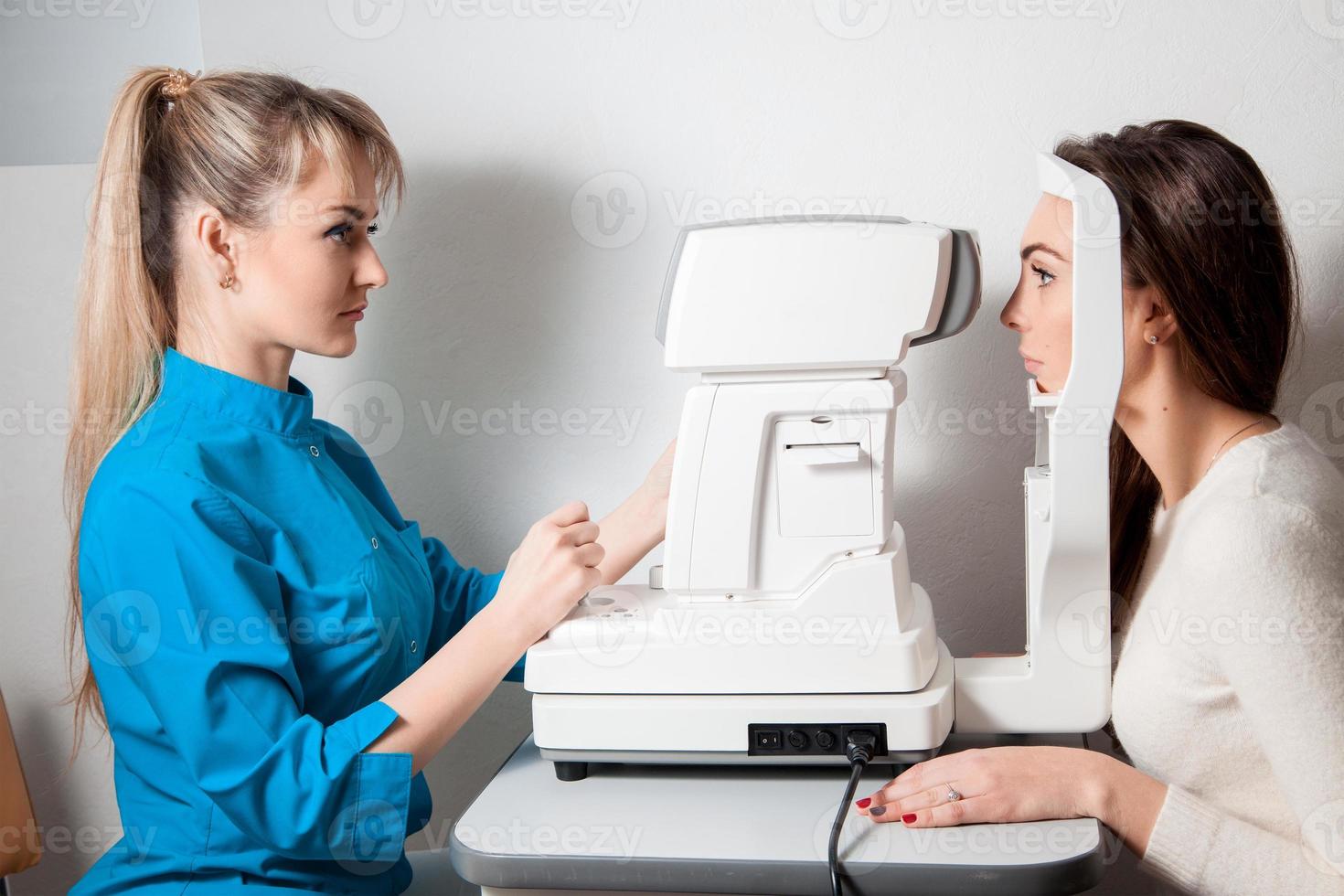 Ophthalmologist woman In Exam Room With Young beautiful brunette Sitting In Chair Looking Into Eye Test Machine photo
