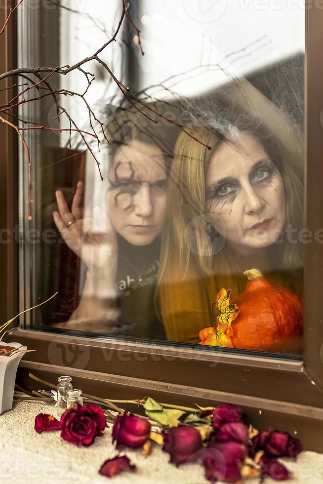 Two female witches look through a spider web ominously out of the window against the backdrop of Halloween decorations. Masquerade, Halloween party photo