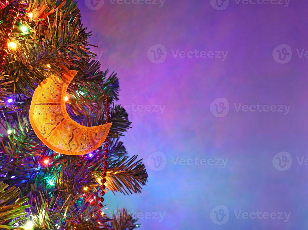 decoración navideña hecha a mano luna amarilla con adorno en abeto en la noche festiva, guirnalda de colores, cuentas rojas, fondo púrpura borroso. tarjeta de felicitación navideña con espacio de copia. foto