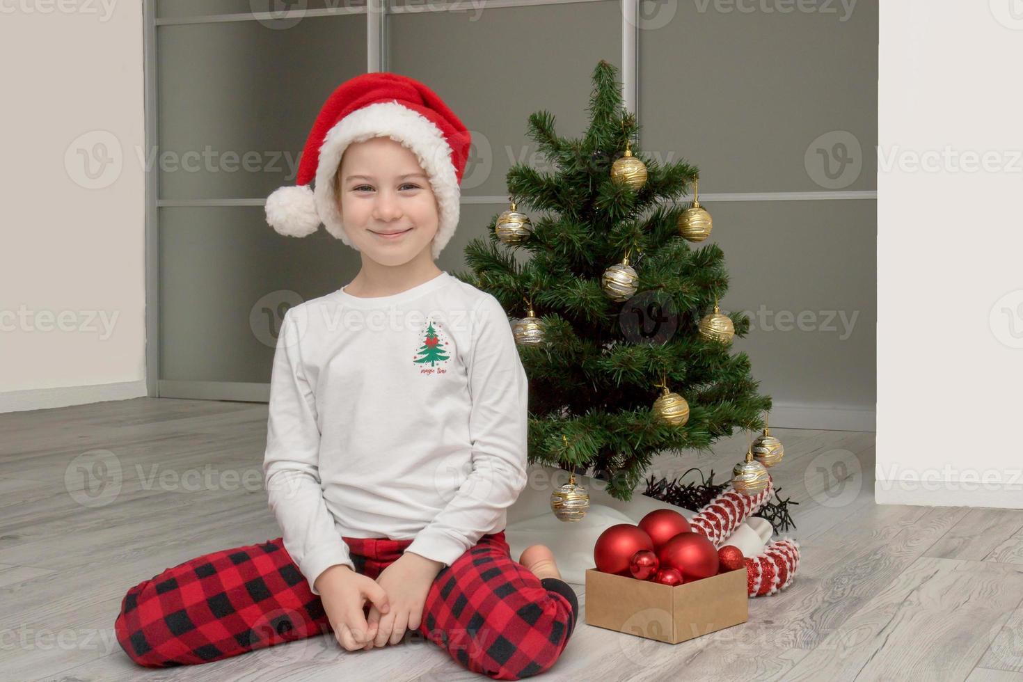 girl in plaid pajamas and a santa hat sits next to the Christmas tree and smiles, Christmas photo