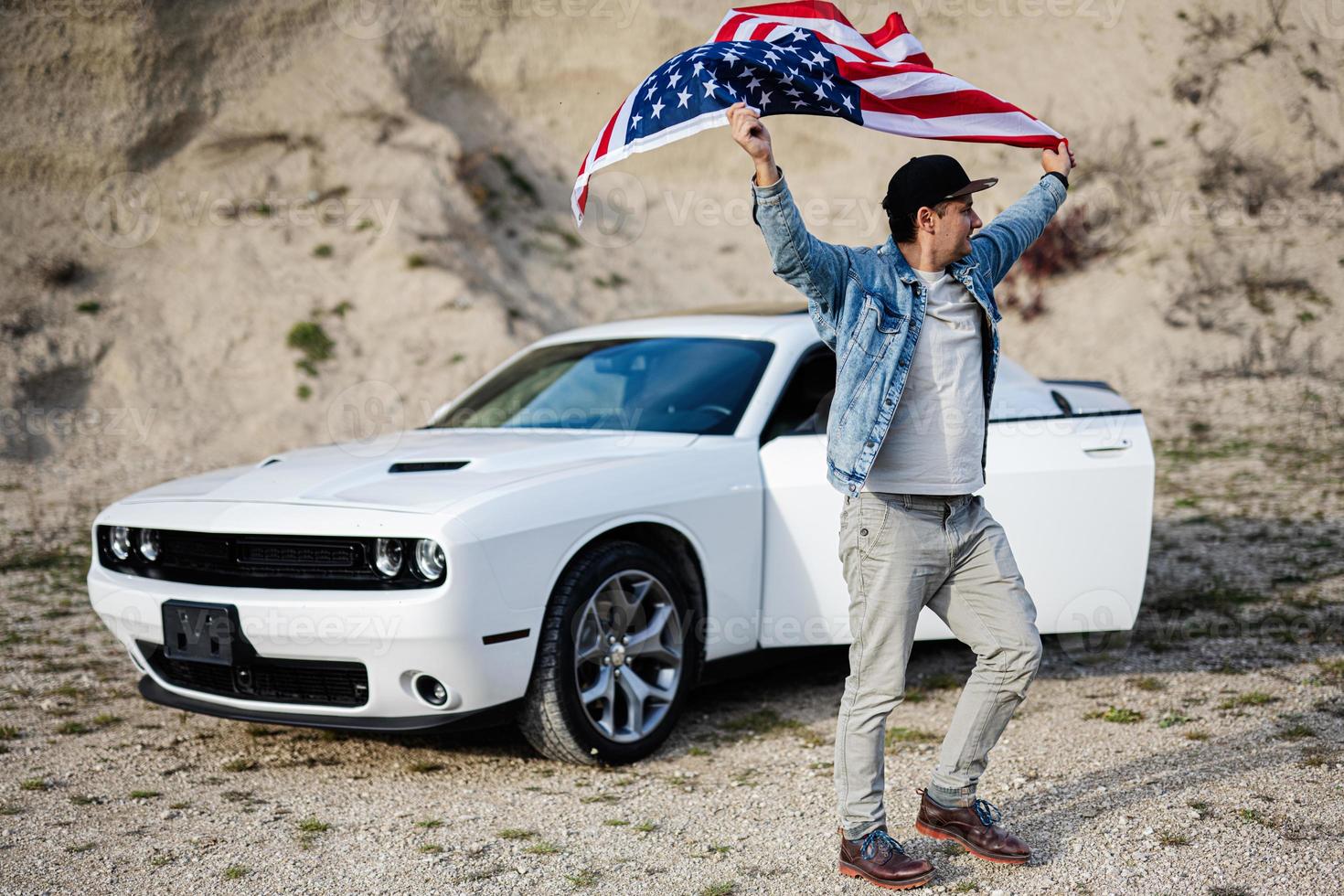 Handsome man in jeans jacket and cap with USA flag near his white american muscle car in career. photo
