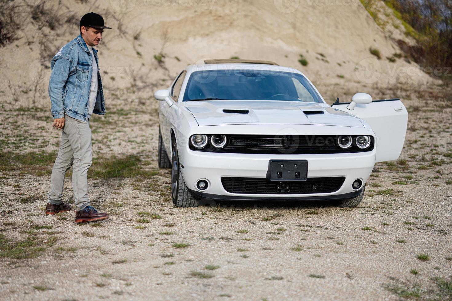 un hombre guapo con chaqueta de jeans y gorra está parado cerca de su auto blanco en carrera. foto