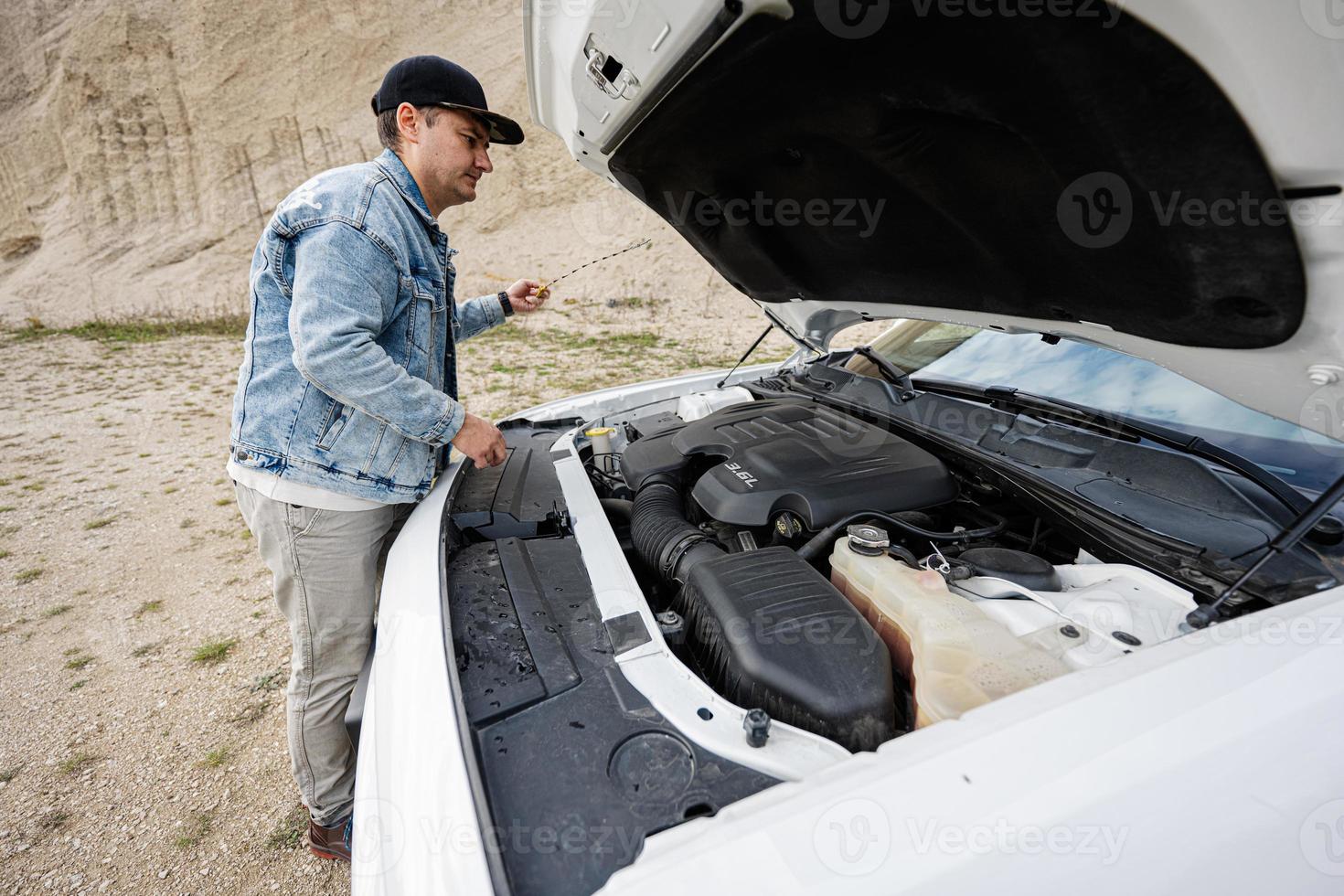 Handsome man in jeans jacket and cap is standing near his white muscle car with opening hood, check oil level of engine. photo