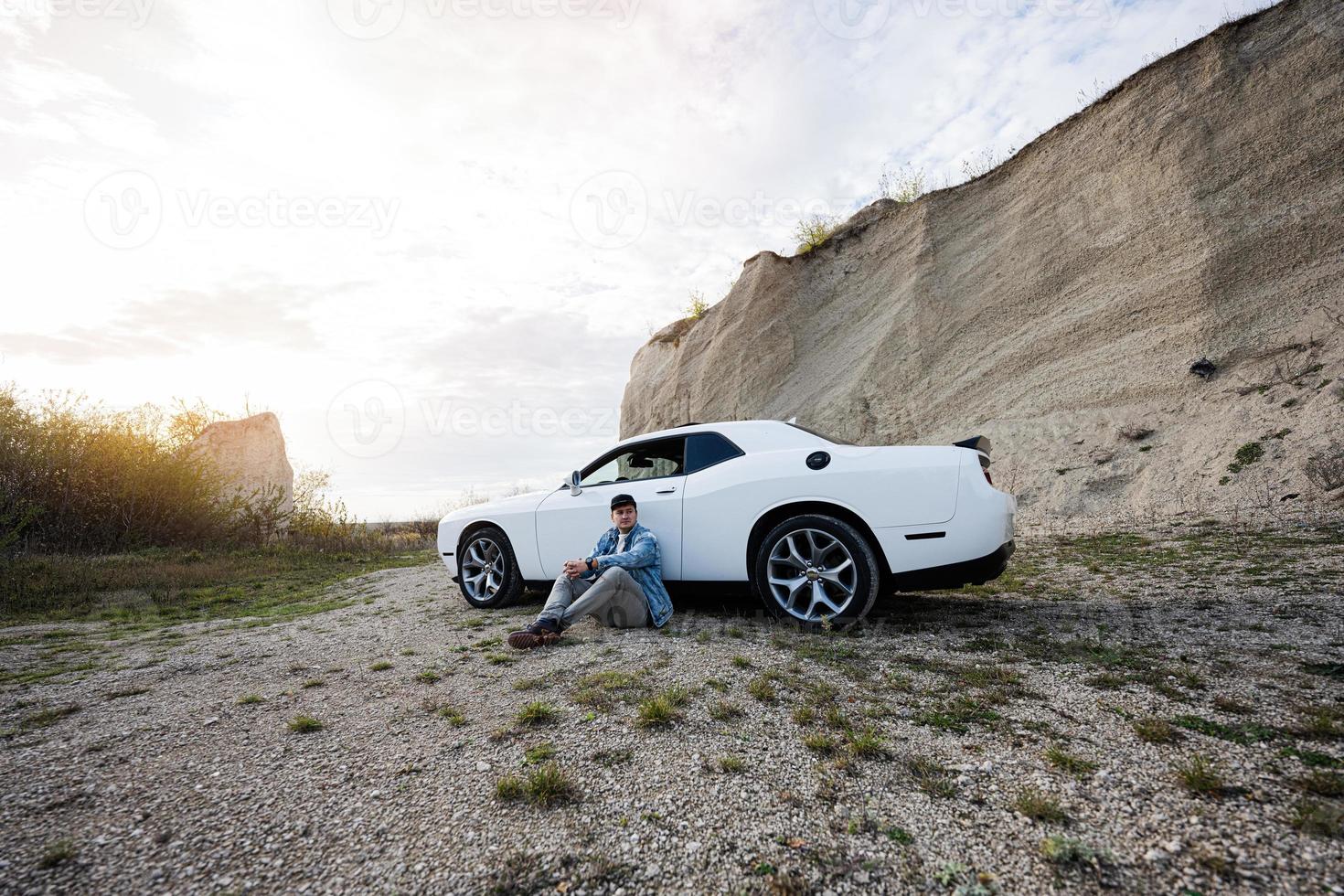 Handsome man in jeans jacket and cap sitting near his white muscle car in career. photo