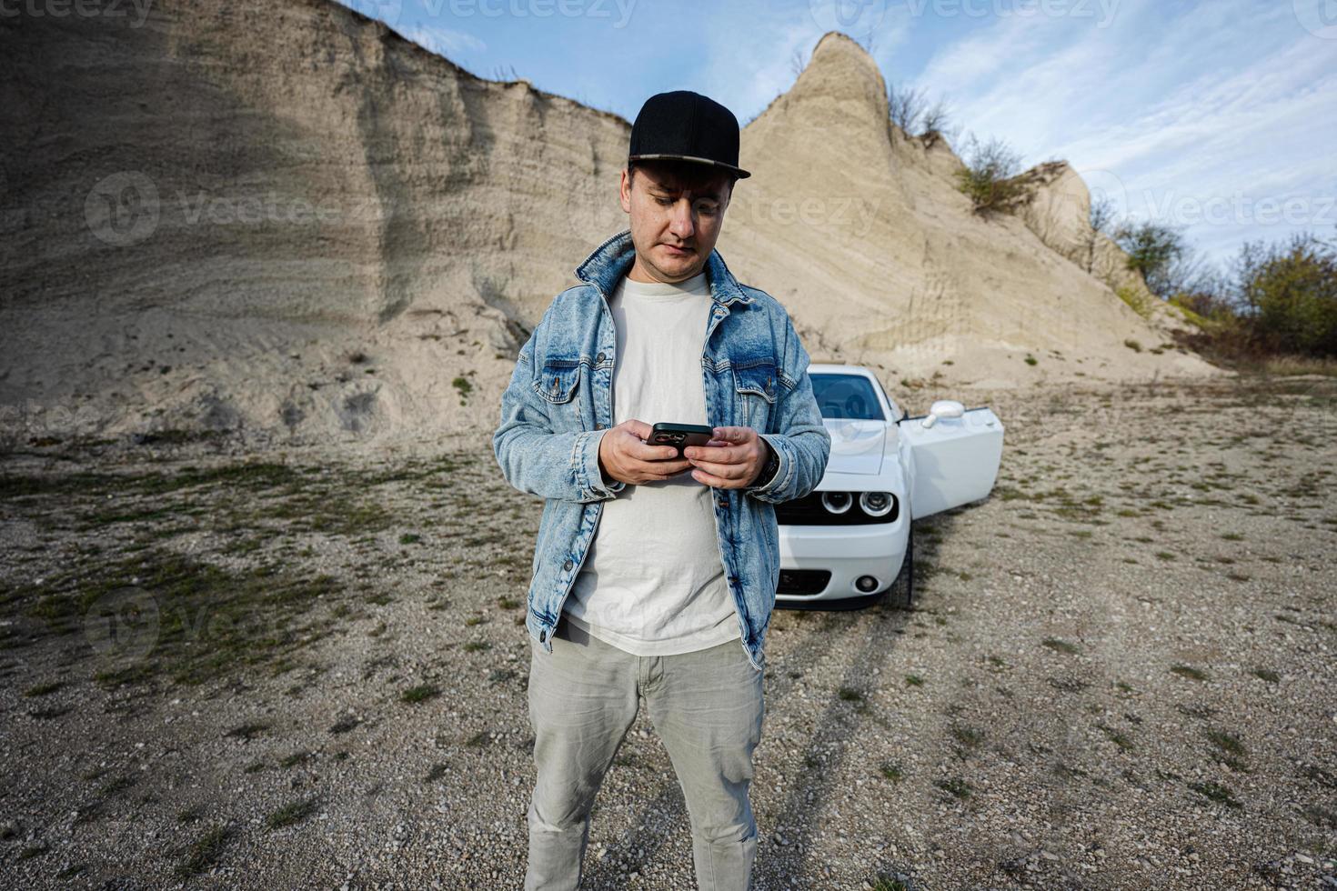 Handsome man with mobile phone, wear in jeans jacket and cap, standing near his white muscle car in career. photo