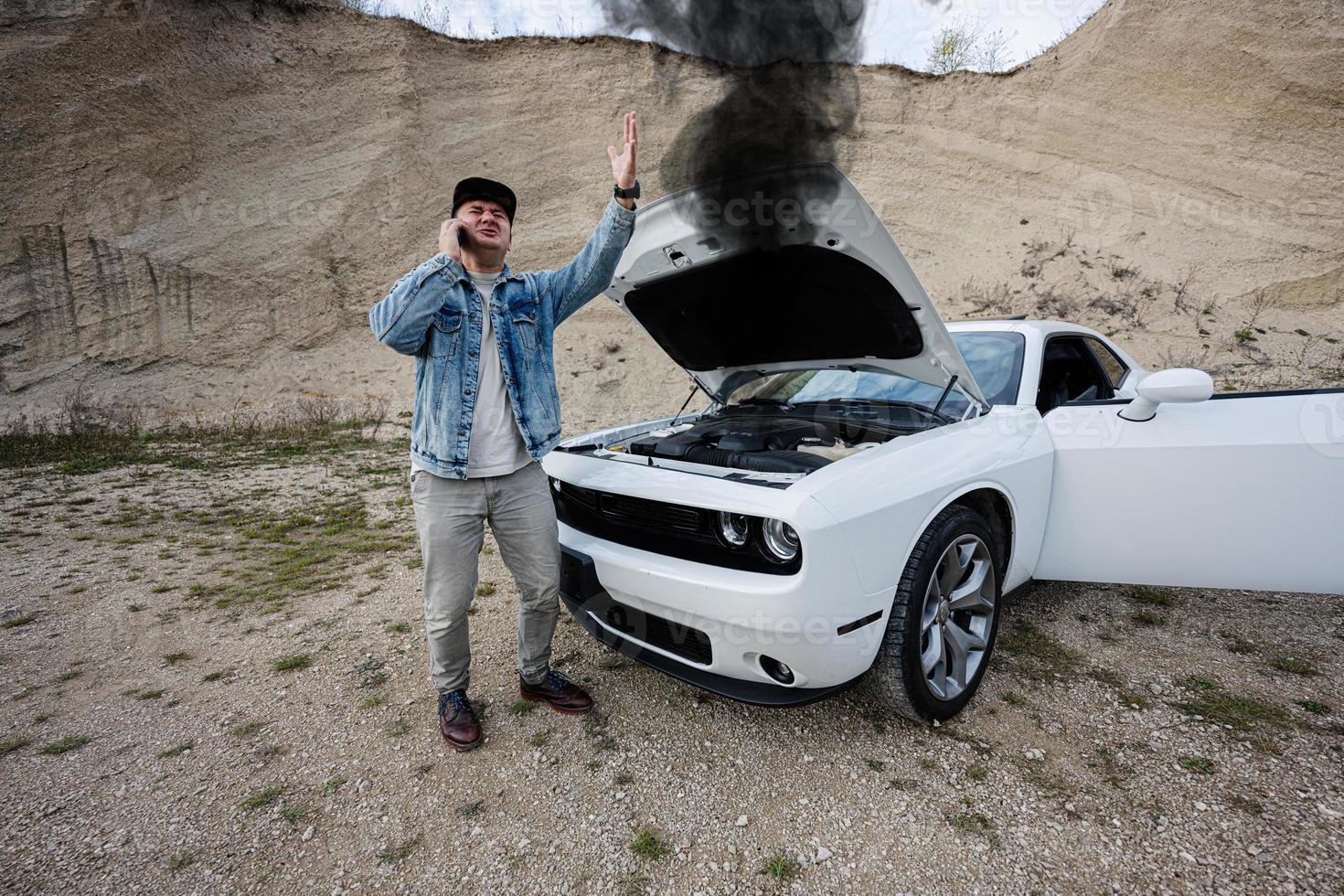 Shocked man calling on phoen to emergency near  his broken muscle car, open hood, smoke from engine,  breakdown, need help. photo
