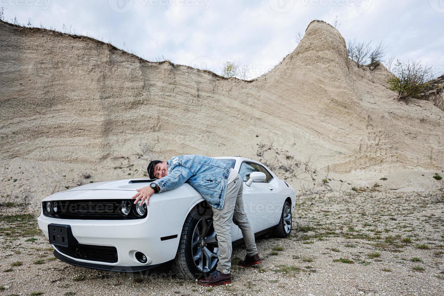 feliz nuevo dueño hombre en chaqueta de jeans y gorra está de pie cerca de su sueño - coche de músculo blanco. foto