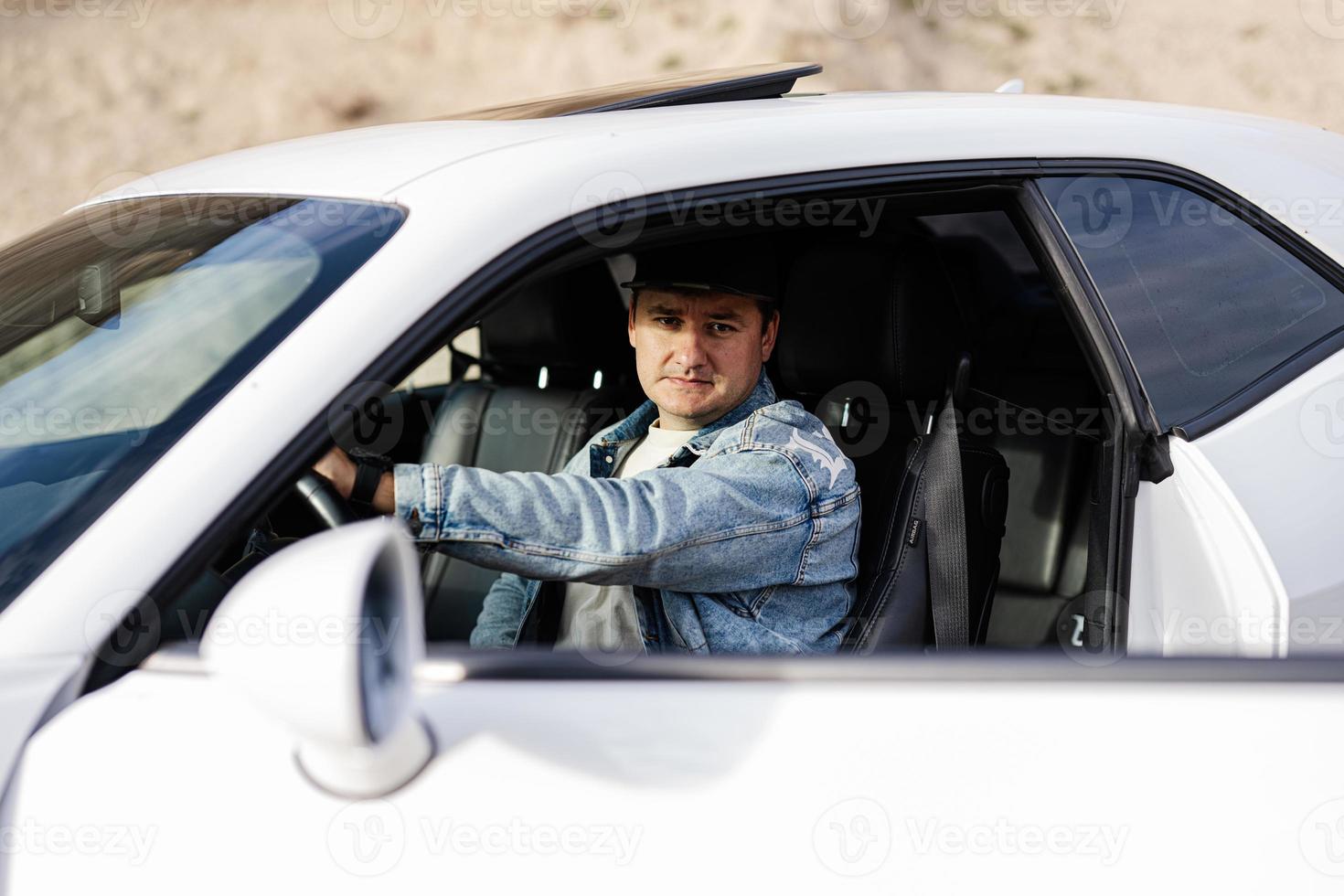 Handsome man in jeans jacket and cap sit at his white muscle car. photo