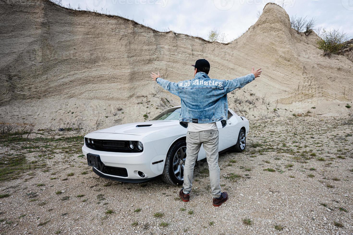 Back of man in jeans jacket and cap is standing and hands to the sides, near his white muscle car in career. Los Angeles. photo