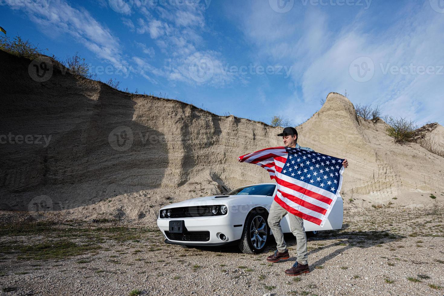 Handsome man in jeans jacket and cap with USA flag near his white american muscle car in career. photo