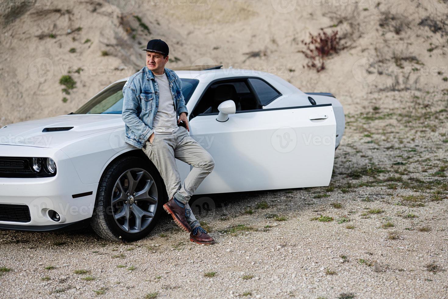 Handsome man in jeans jacket and cap is standing near his white muscle car in career. photo