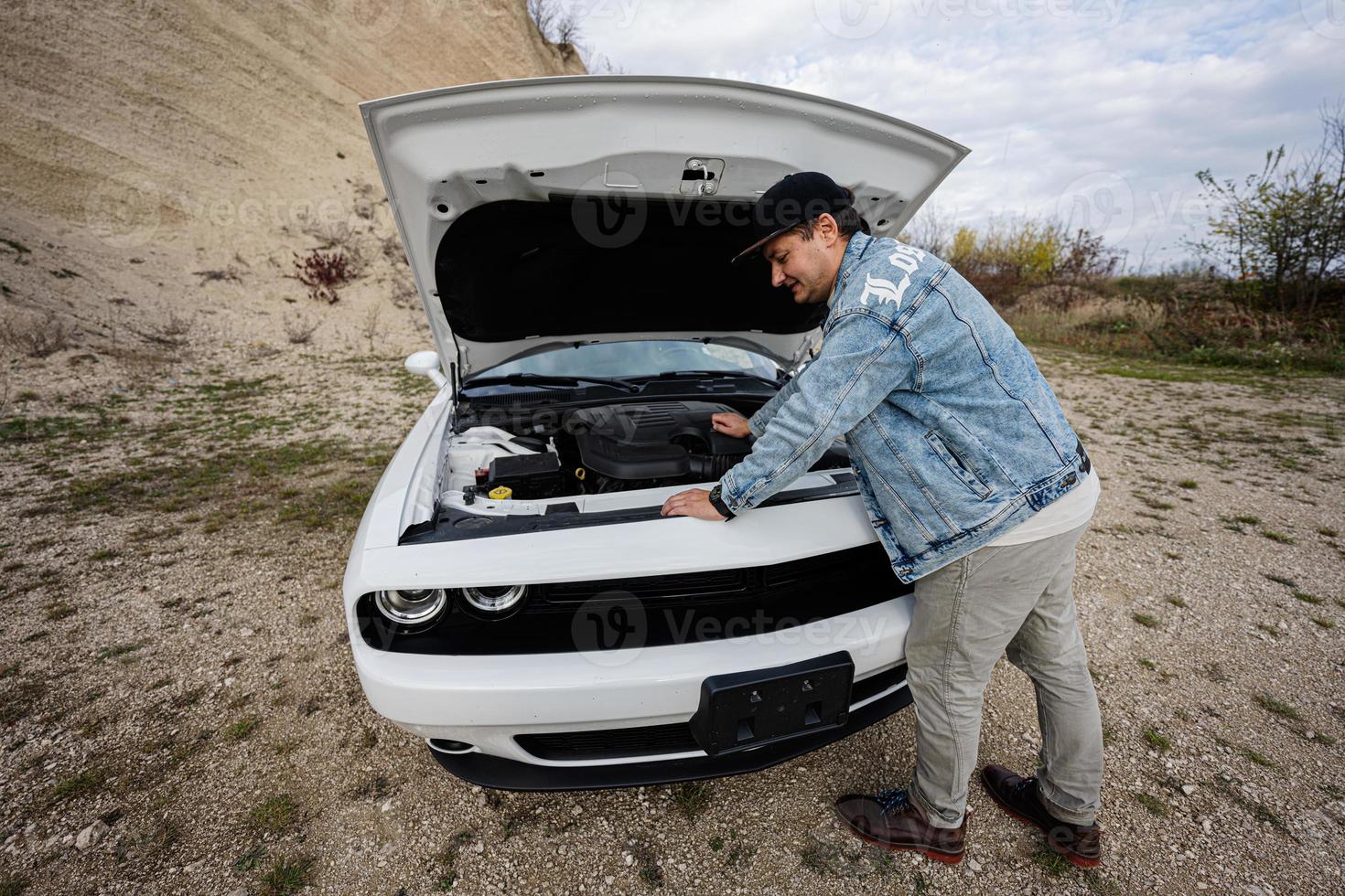 Handsome man in jeans jacket and cap is standing near his white muscle car with opening hood, check engine. photo