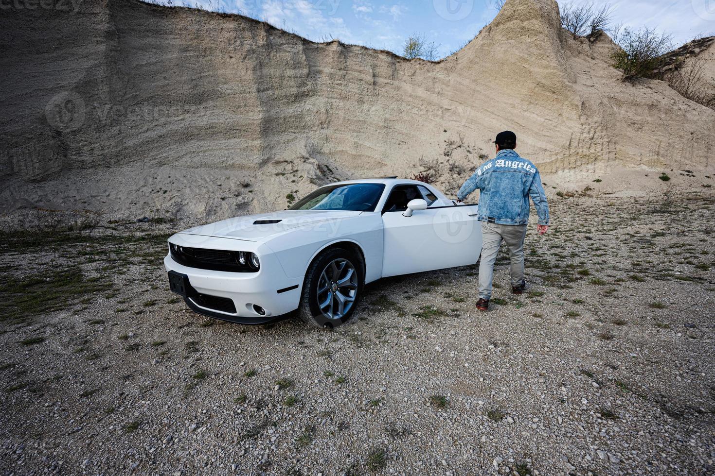 Back of man in jeans jacket and cap open door of his white muscle car in career. Los Angeles. photo