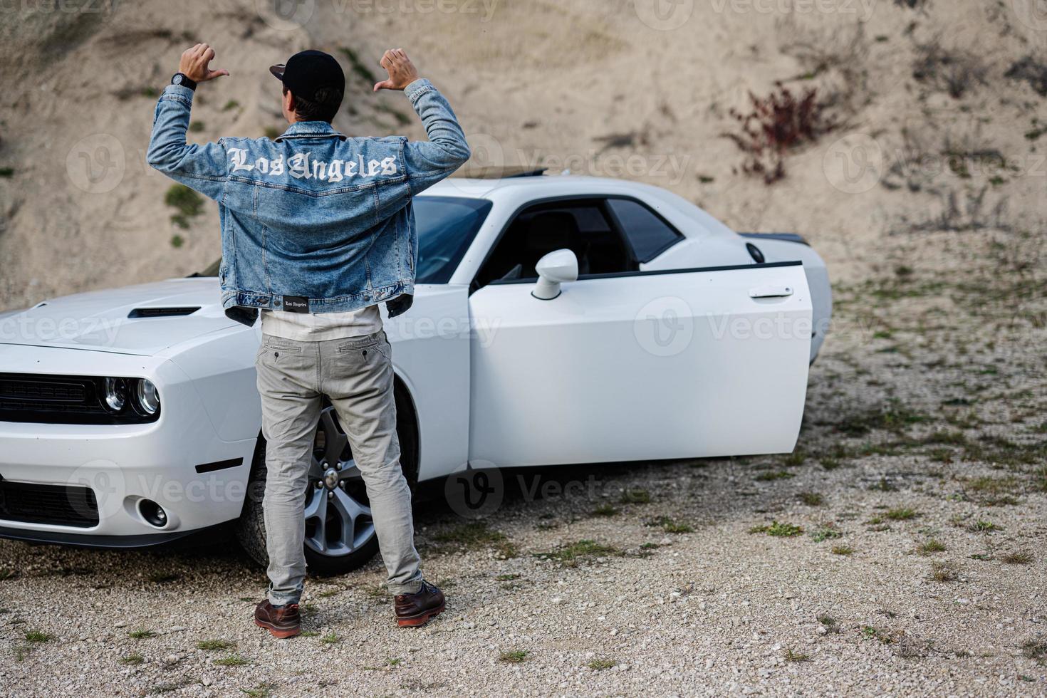 Back of man in jeans jacket and cap is standing , near his white muscle car in career. Los Angeles. photo