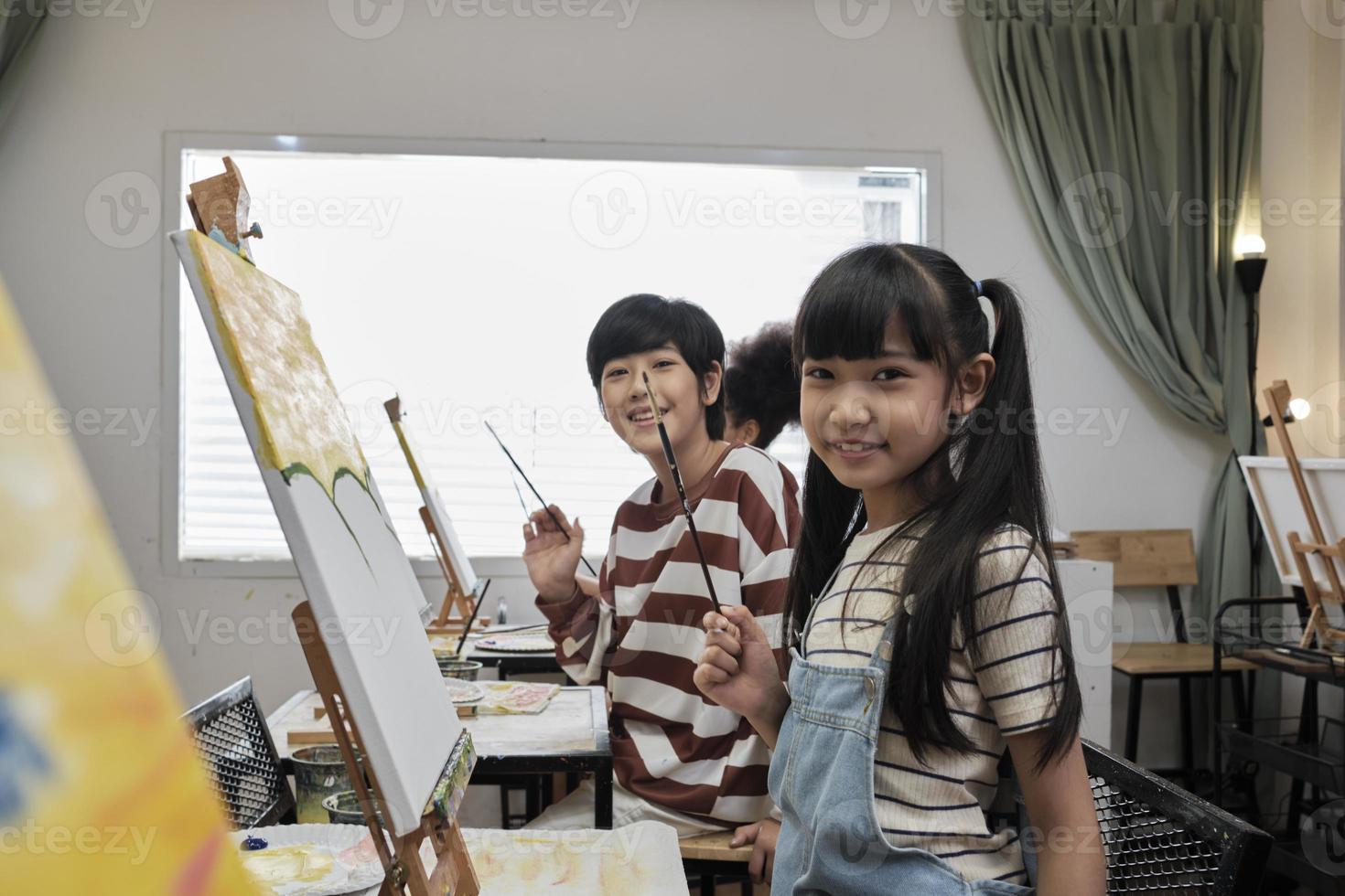 Portrait of Asian kids looking at camera and smiling with acrylic color picture painted on canvas in art classroom and creative learning with talents and skill at elementary school studio education. photo