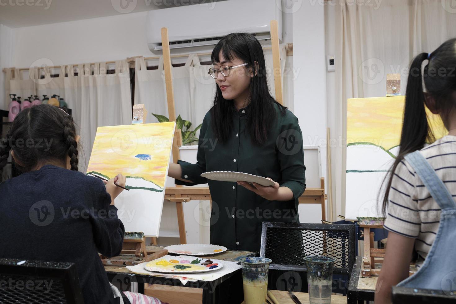A female Asian teacher teaches and demonstrates to the children on acrylic color picture painting on canvas in art classroom, creatively learning with skill at the elementary school studio education. photo