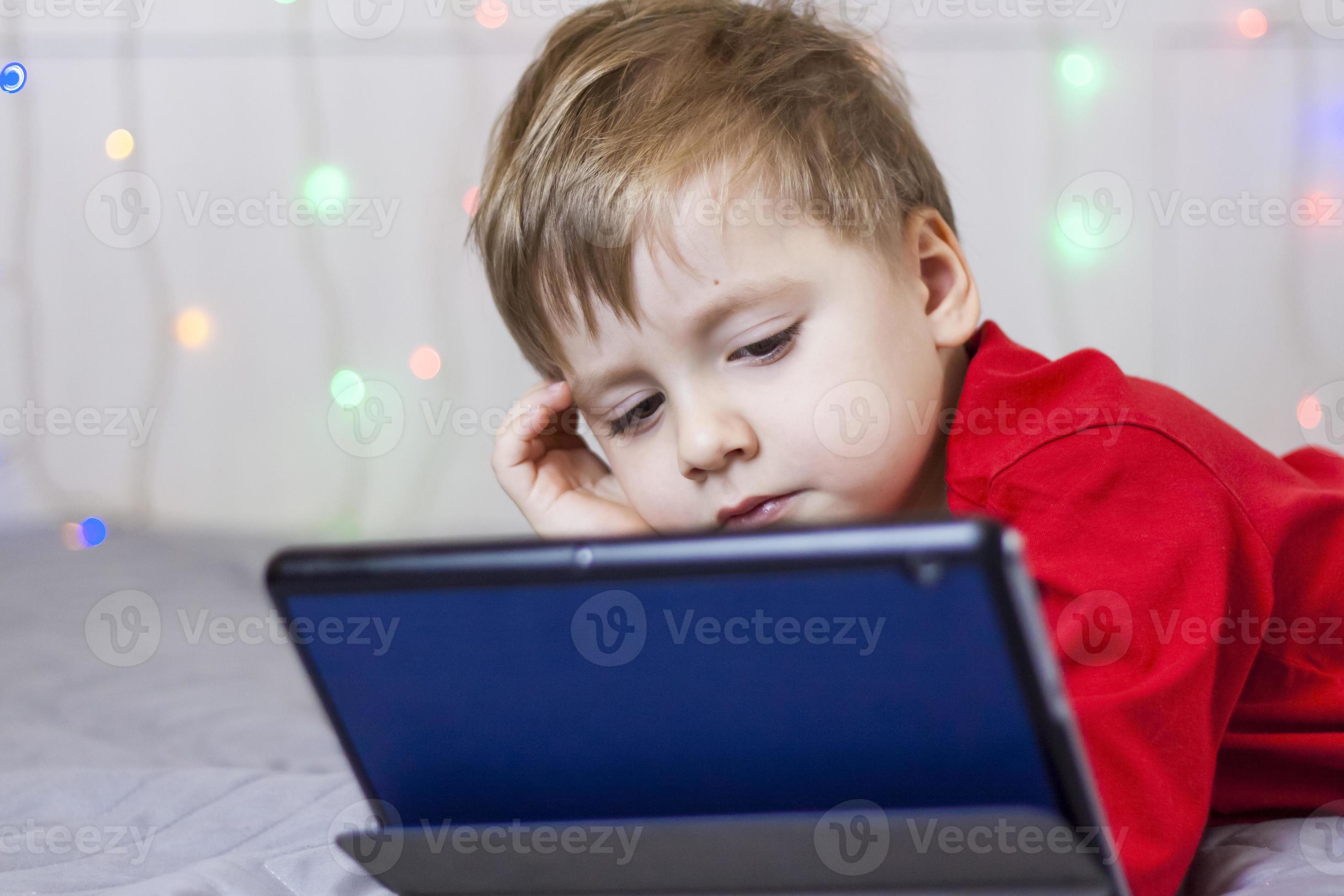 A cute boy is sitting on the bed and looking at a tablet. Funny smiling