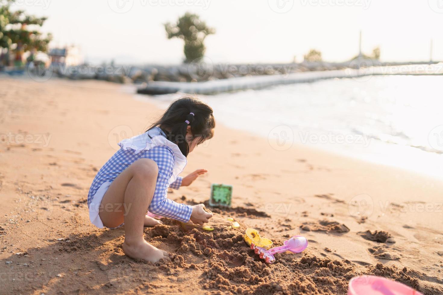 linda niña asiática jugando o haciendo castillos de arena o cavando con arena en una playa tropical. niños con hermoso mar, arena y cielo azul. niños felices de vacaciones en la playa corriendo en la playa. foto