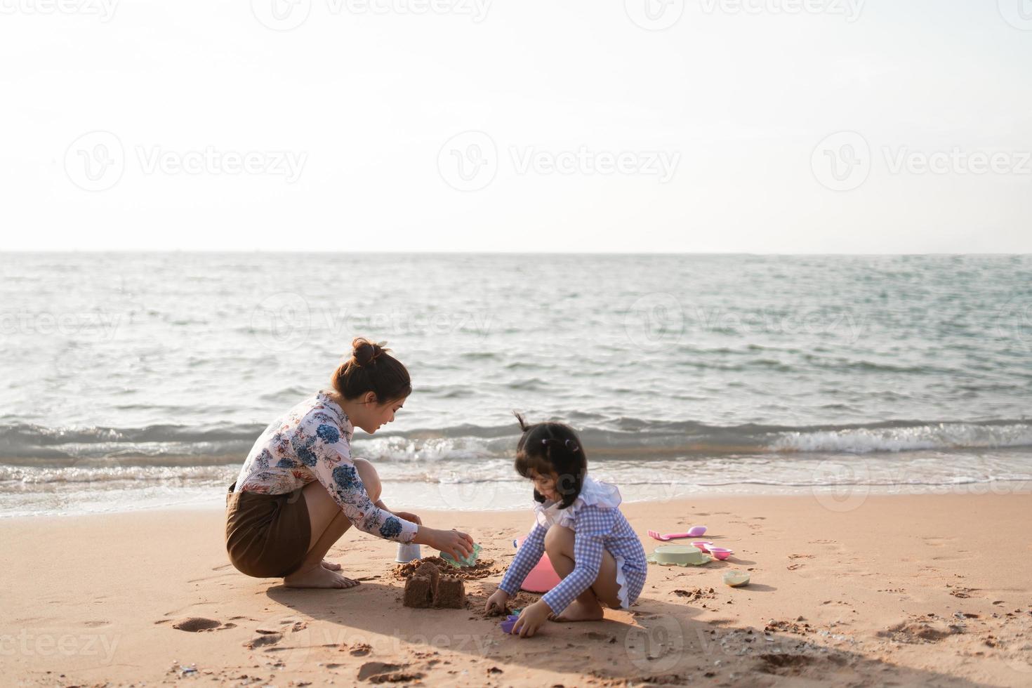 Asian cute little girl and her mother playing or making sand castle or digging with sand on tropical beach. Children with beautiful sea, sand blue sky. Happy kids on vacations seaside on the beach. photo