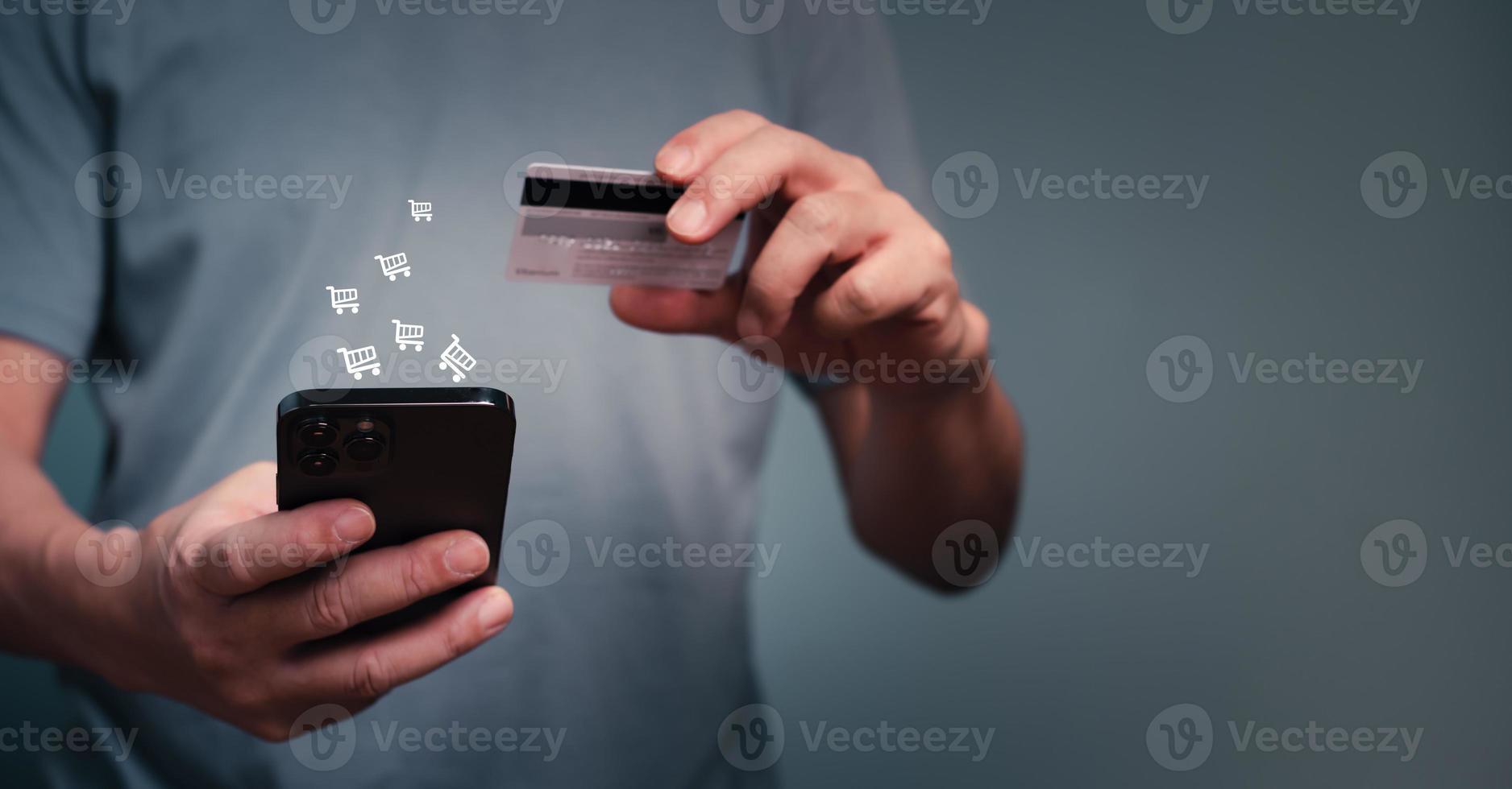 Young man using mobile phone and credit card for online shopping, entering number to make payment, buying items, holding the debit card for internet banking account with cart icons. photo