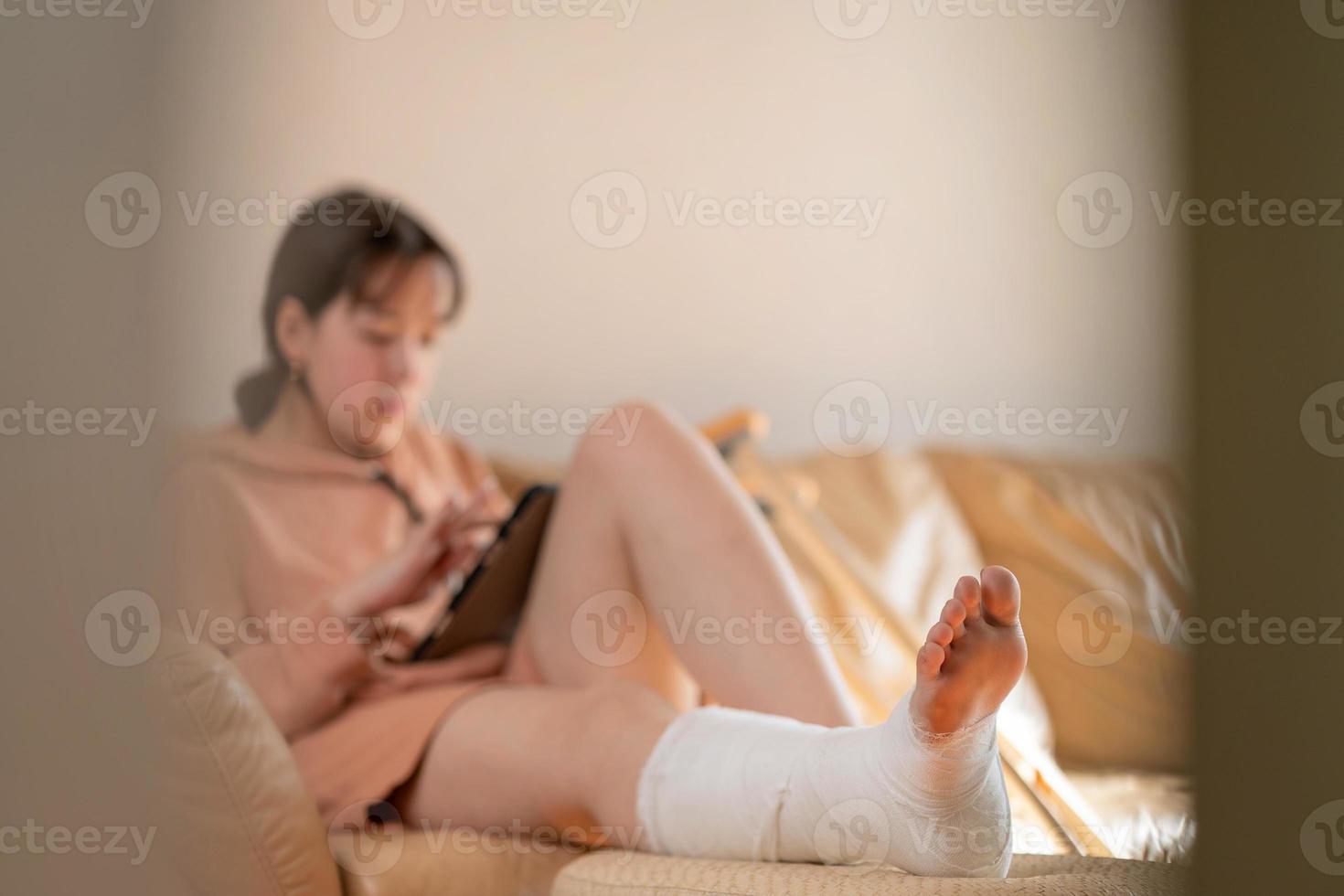 Girl with a cast on their leg, sitting on the couch and drawing on a graphics tablet. Bandaged leg cast and toes after a running injury accident.Teen girl in a plaster cast. photo