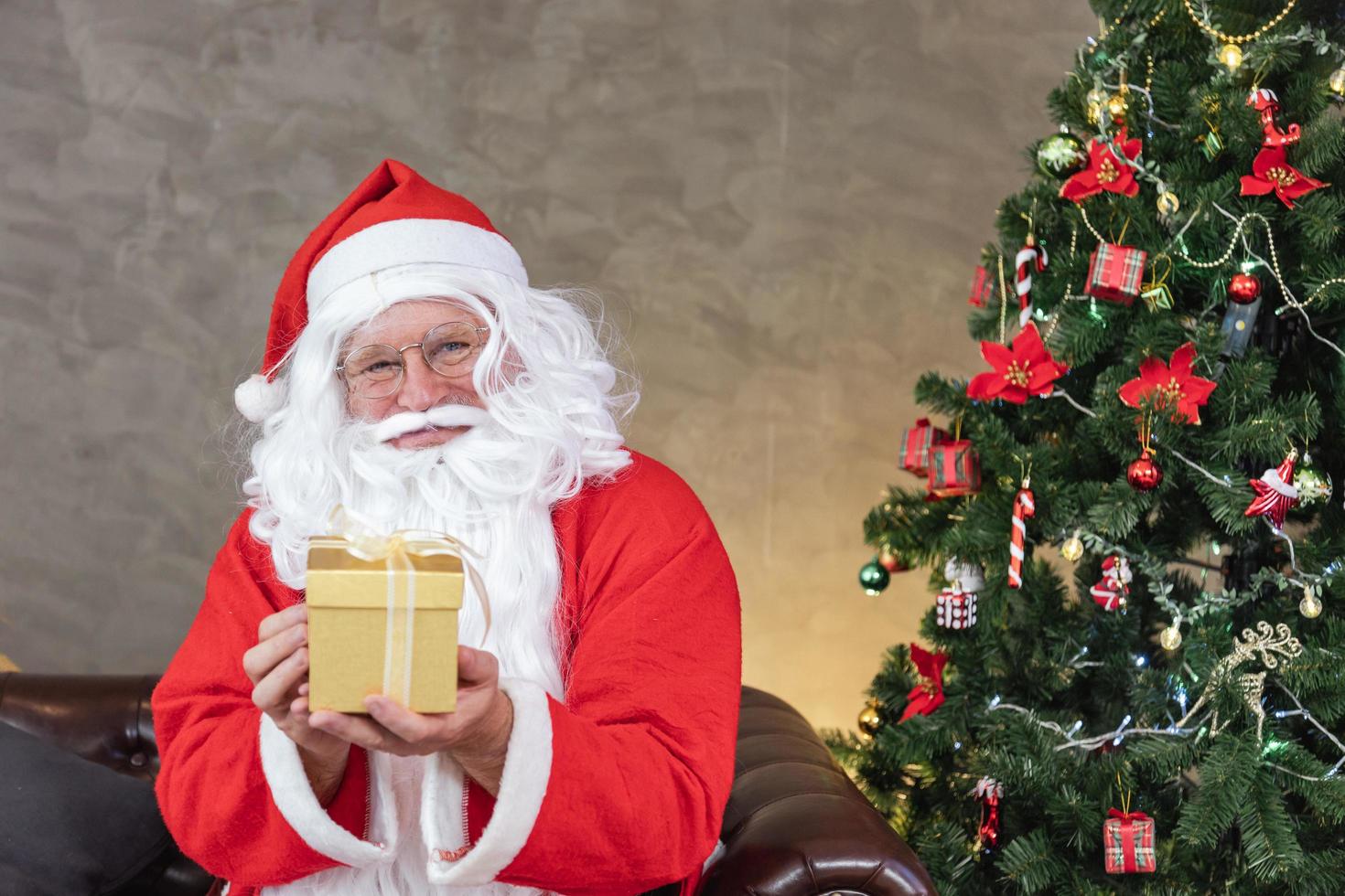 Santa Claus is holding christmas present box with fully decorated chrsitmas tree for season celebration and happy new year event photo