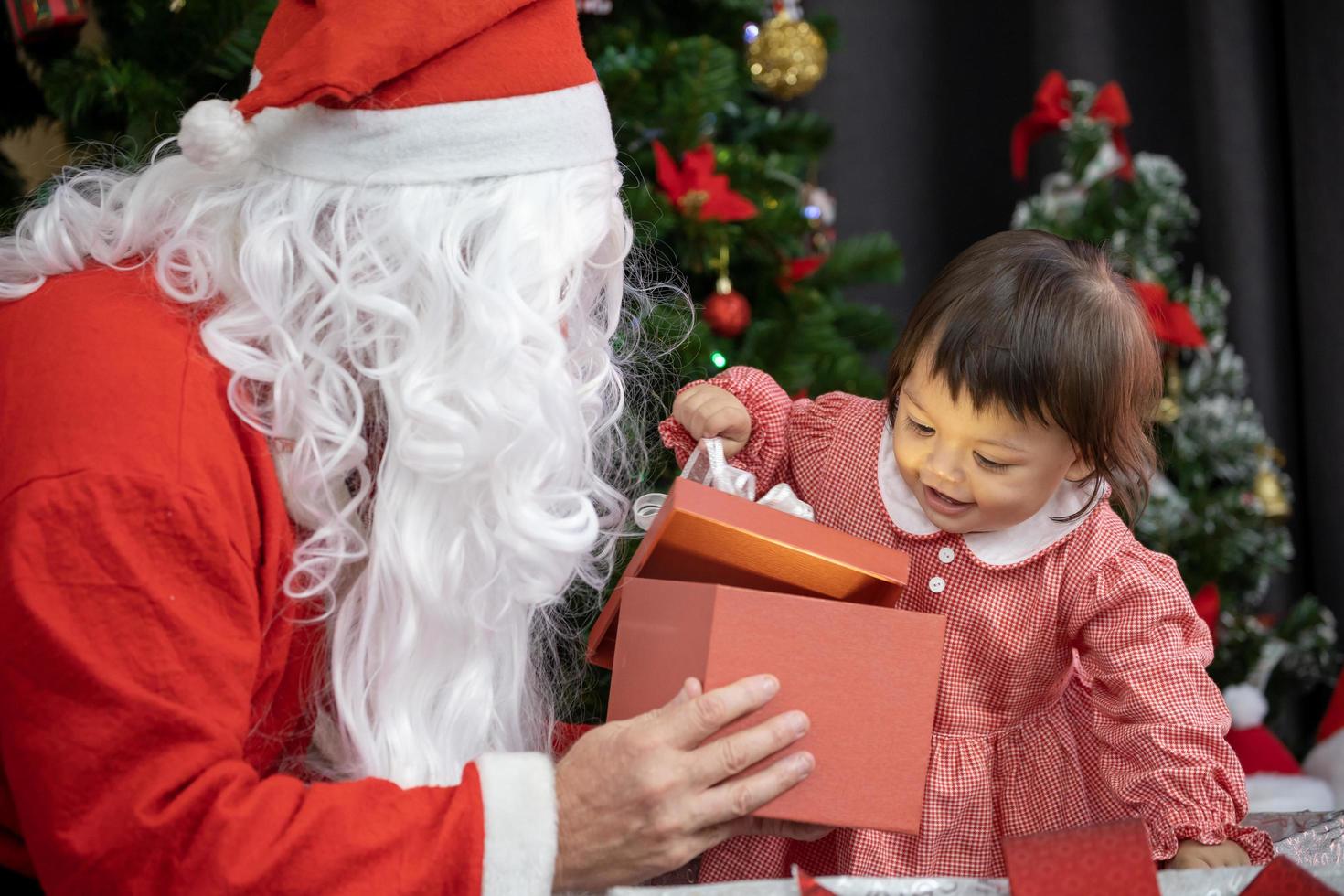 el bebé caucásico está recibiendo regalos de santa claus por la noche junto al árbol de navidad completamente decorado para el concepto de celebración de temporada foto