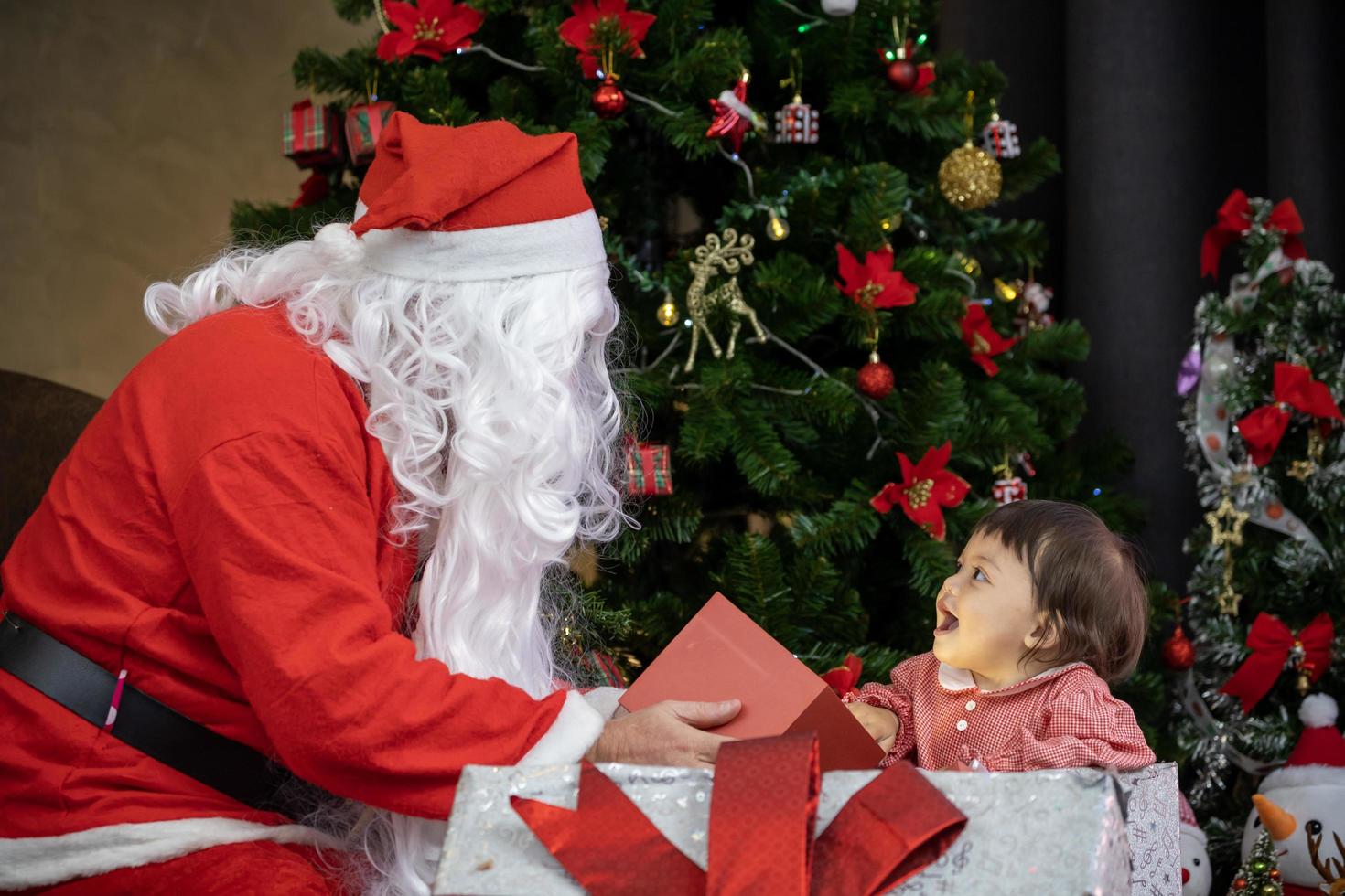 Caucasian baby is getting present from Santa claus at night by the fully decorated christmas tree for season celebration usage photo