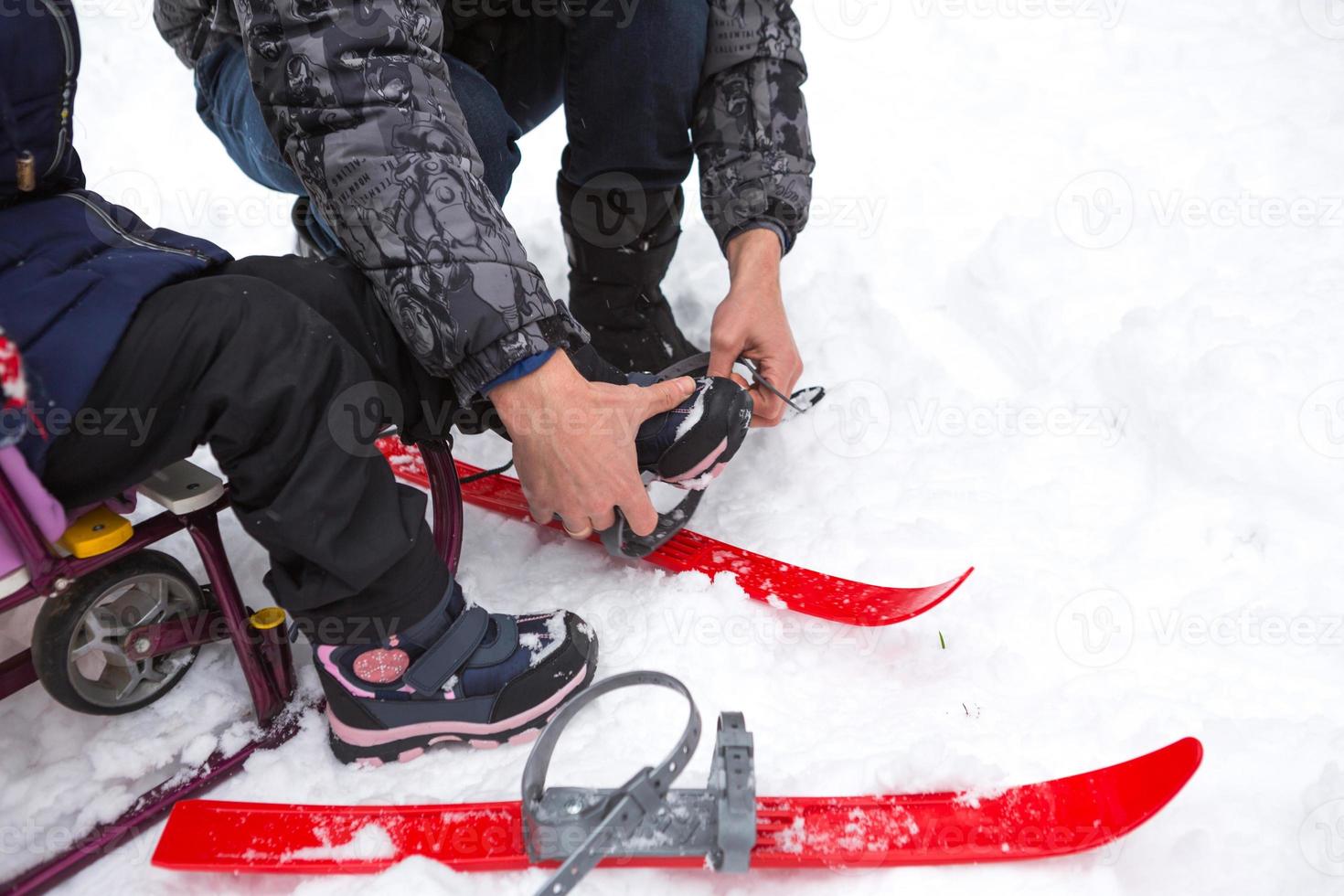 papá ayuda a su pequeña hija a ponerse esquís de plástico para niños. entrenamiento deportivo, esquí, apoyo y asistencia. deportes activos de invierno al aire libre desde la infancia foto