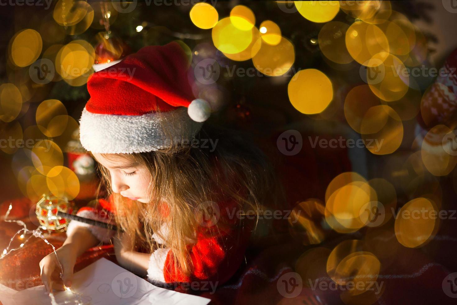 Little girl in a Santa hat and red dress under Christmas tree is dreaming, waiting for the holiday, lying on a plaid blanket. A letter on piece of paper, gifts. New year, Christmas. defocus lights photo