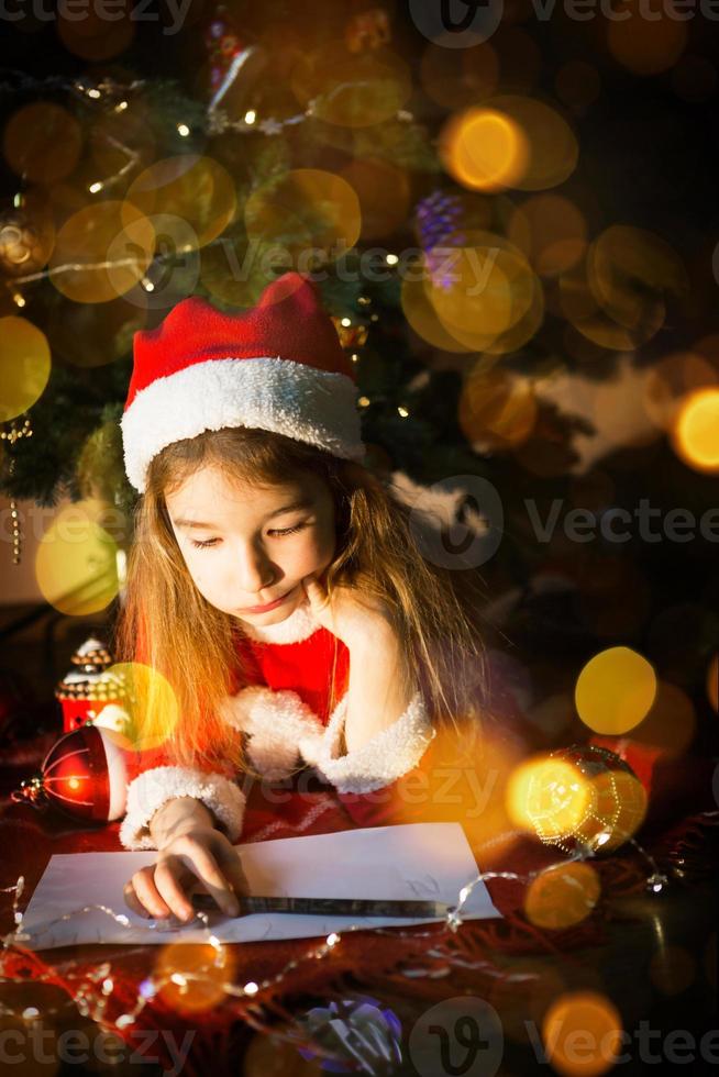 Little girl in a Santa hat and red dress under Christmas tree is dreaming, waiting for the holiday, lying on a plaid blanket. A letter on piece of paper, gifts. New year, Christmas. defocus lights photo