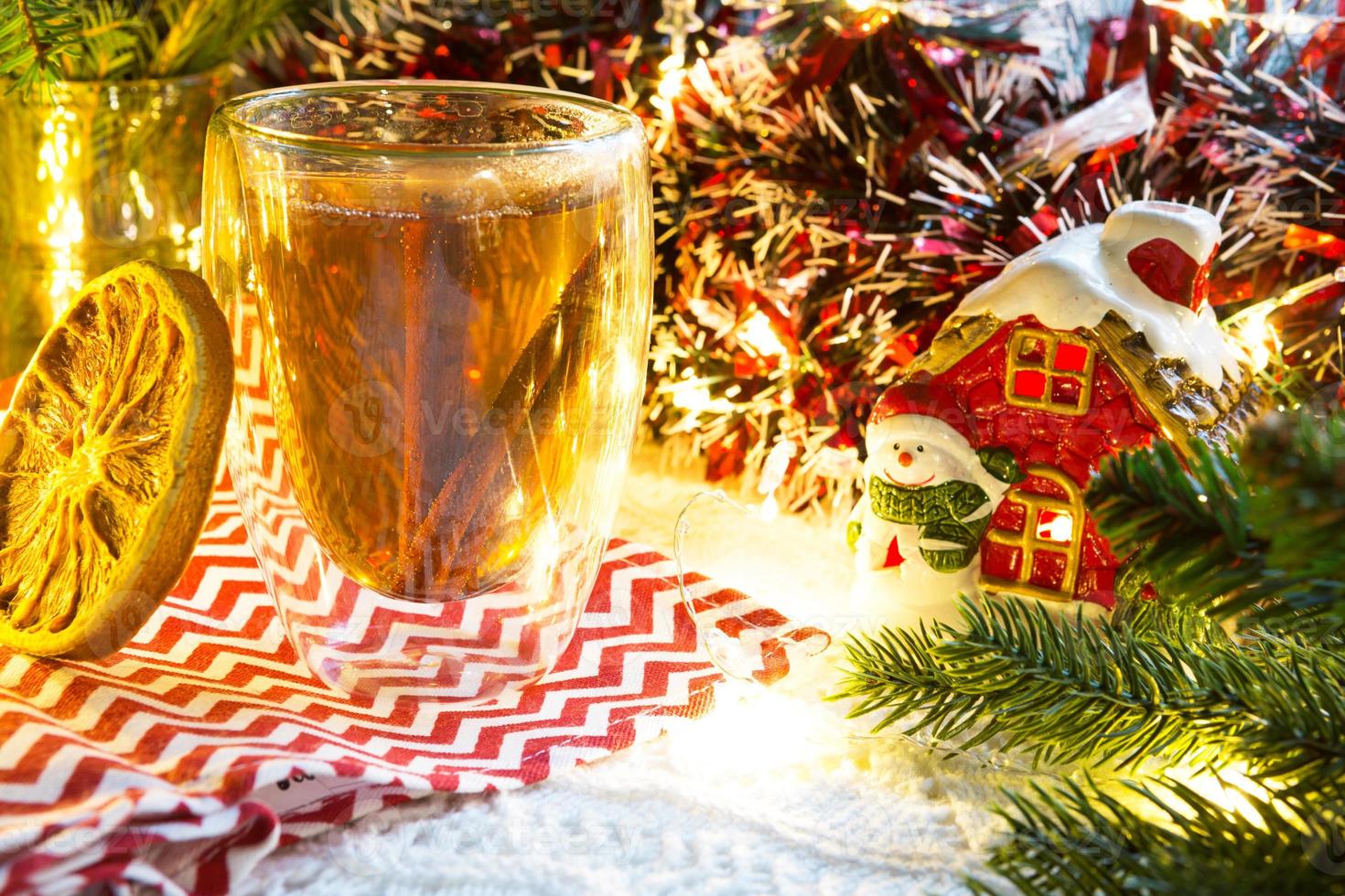 Transparent double-walled glass tumbler with hot tea and cinnamon sticks on the table with Christmas decor and small house. New year's atmosphere, slice of dried orange, garland, spruce branch, cozy photo
