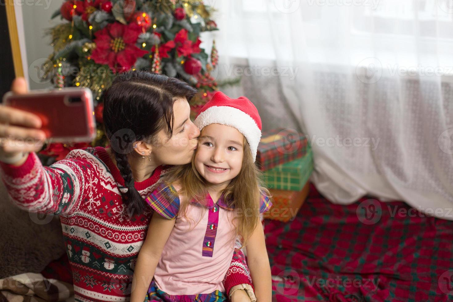 Mom and daughter are sitting near the Christmas tree, hugging, taking photos and selfies on the phone, communicating via video communication, having fun. Christmas, new year's eve at home. Copy space