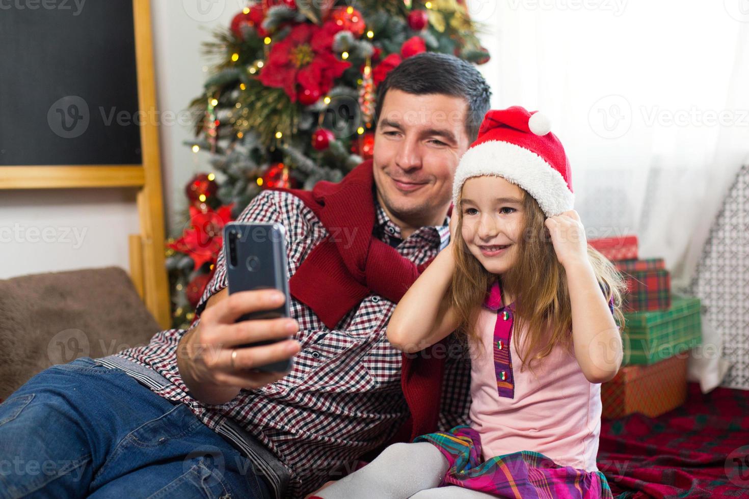 Dad and daughter near the Christmas tree with a smartphone take a selfie, communicate via video connection. Christmas greetings, a gift box, a girl in a Santa hat waving Hello. New year, holiday decor photo