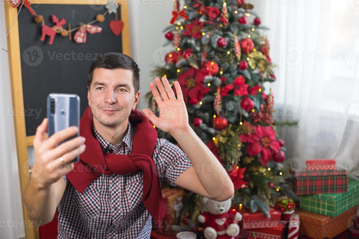 Young man near Christmas tree at home in comfortable clothes waves his hand as sign photo