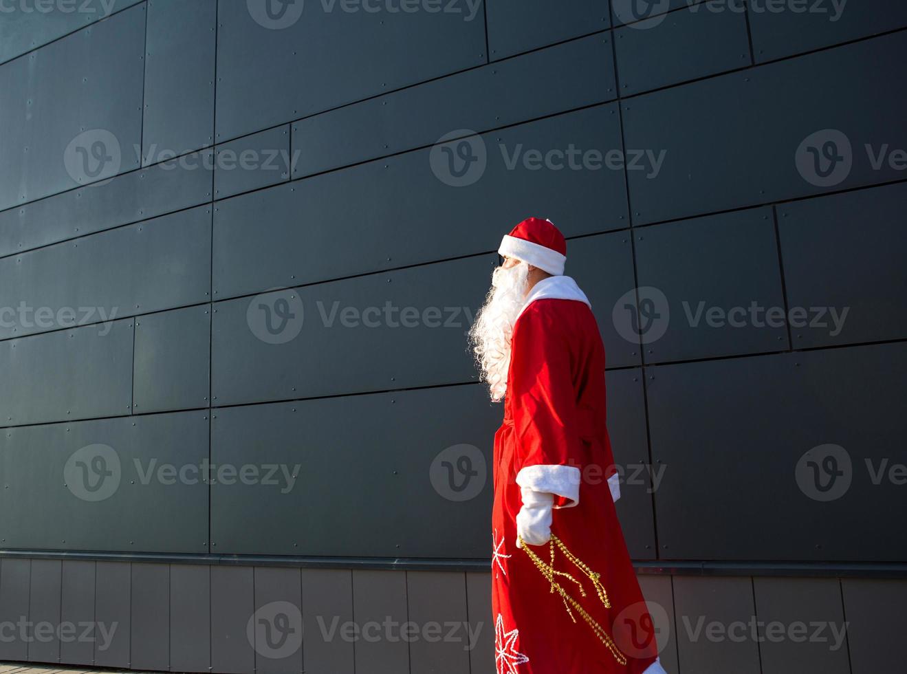 A young man dressed as Santa on the street, standing against a gray modern wall. White beard, a red suit with a hat. Christmas, New year. Russian grandfather Frost. Grey modern background, copy spaces photo