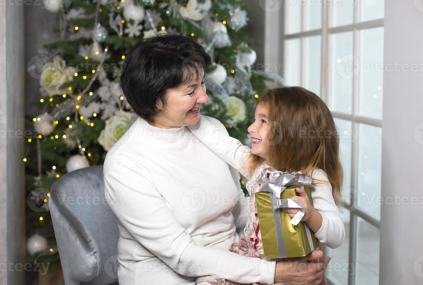 Grandmother with a little girl on the background of Christmas decorations and a large window. Family holiday, emotions, gift box. Granddaughter on granny's lap. New Year photo