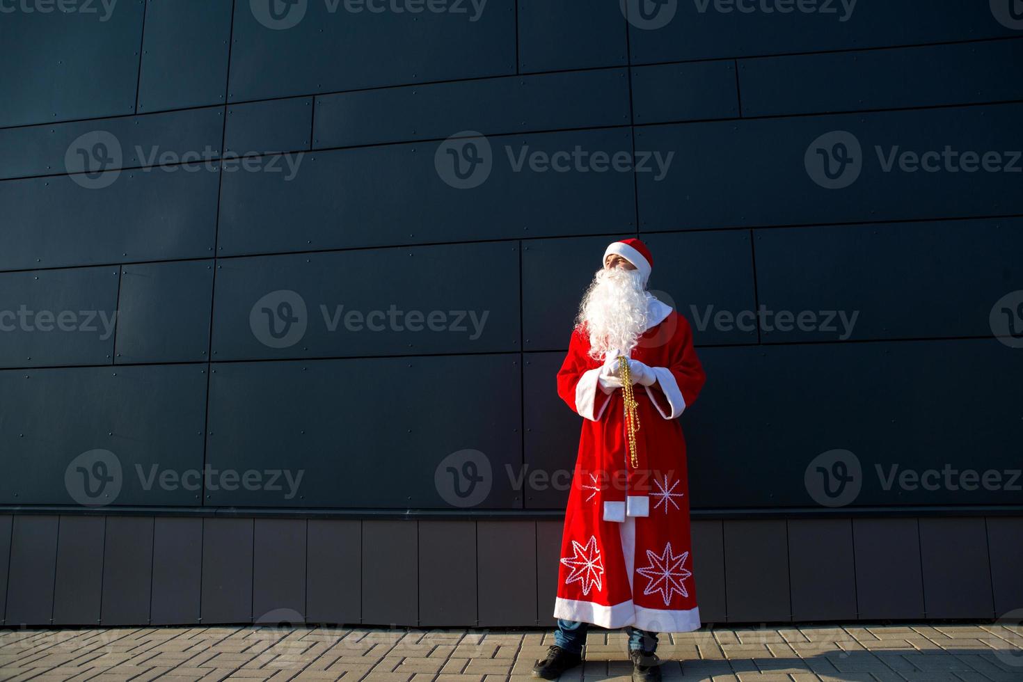 un joven vestido como santa en la calle, parado contra una pared moderna gris. barba blanca, traje rojo con sombrero. navidad, año nuevo. helada del abuelo ruso. fondo moderno gris, espacios de copia foto