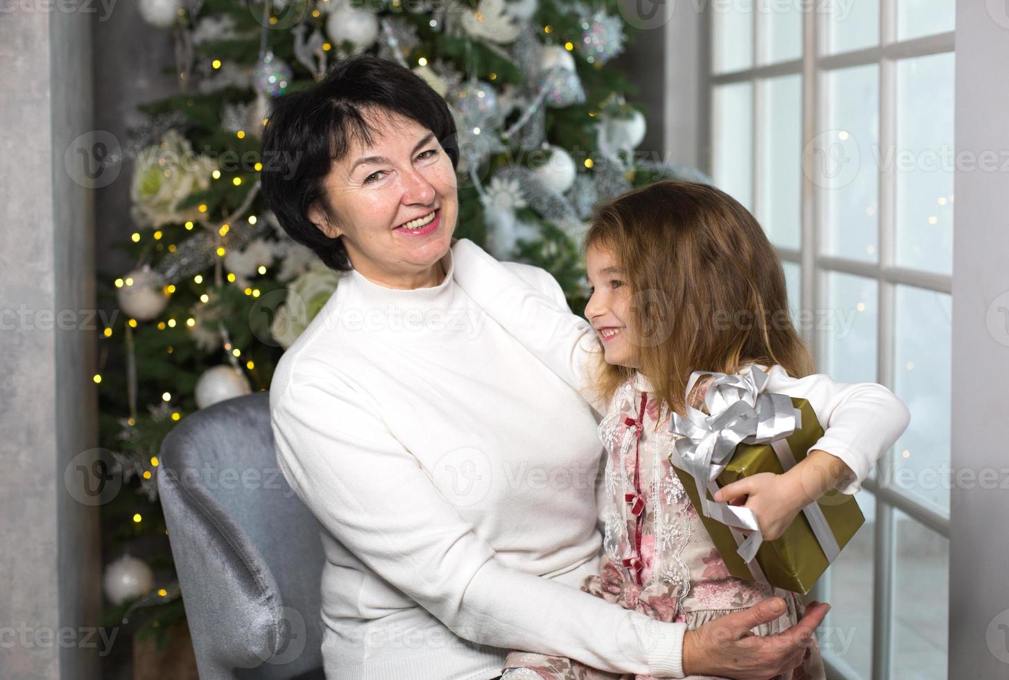 Grandmother with a little girl on the background of Christmas decorations and a large window. Family holiday, emotions, gift box. Granddaughter on granny's lap. New Year photo