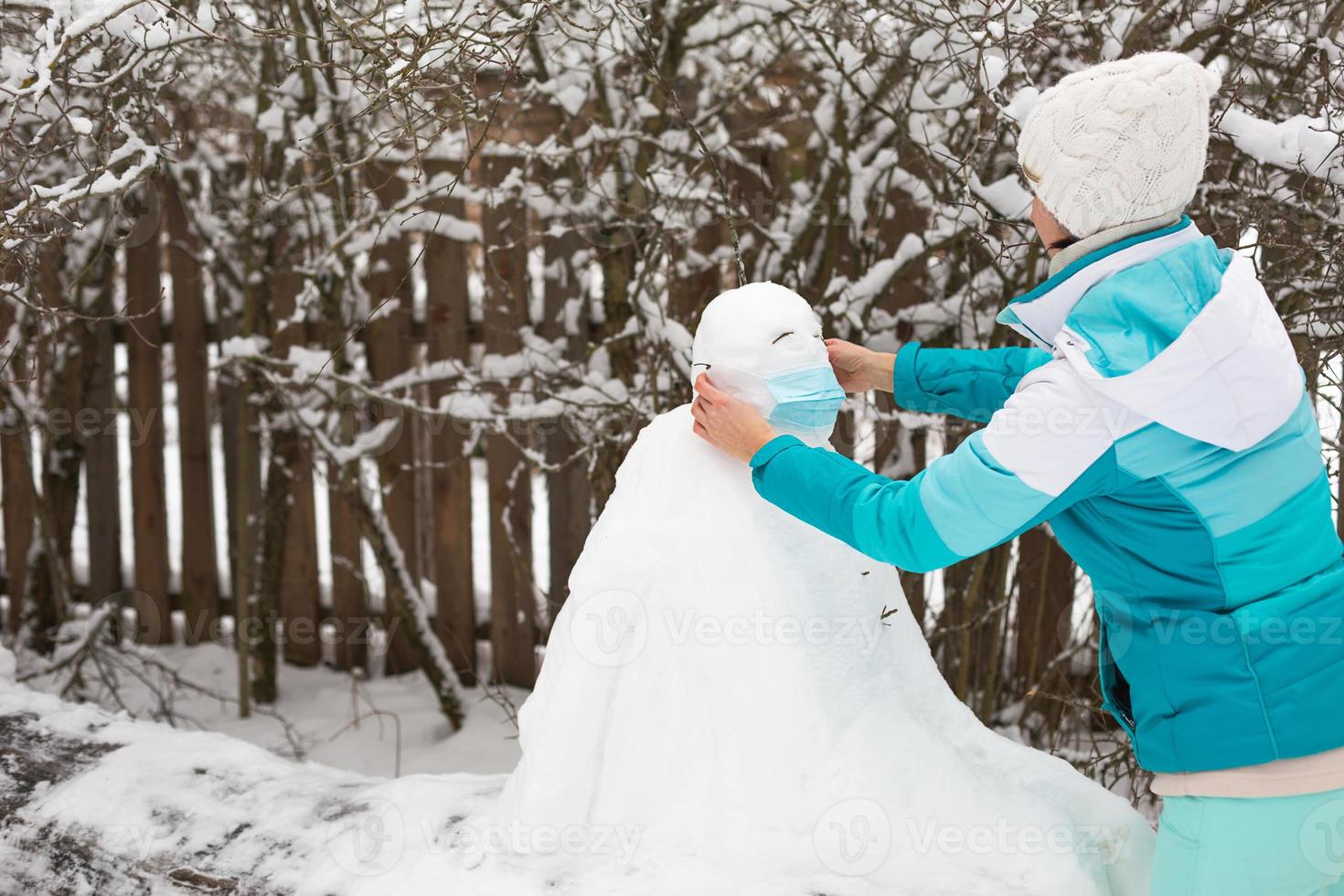 Snowman in a medical mask - a new reality, protection from disease, infection, life in the covid epidemic. Women's hands put on a snowman mask. Winter family outdoor activities photo