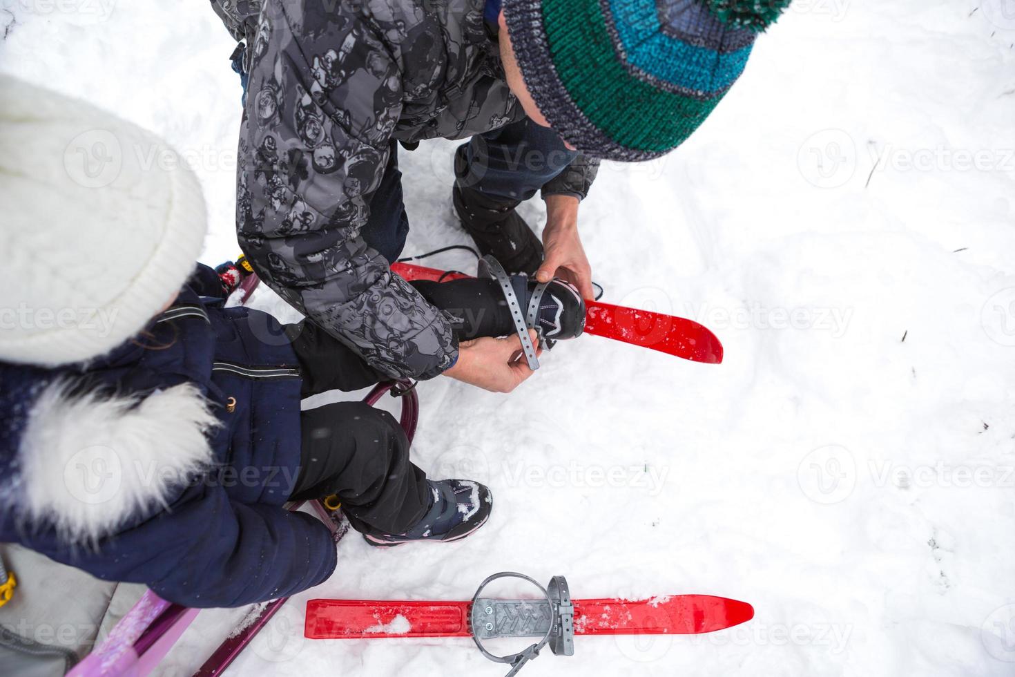 papá ayuda a su pequeña hija a ponerse esquís de plástico para niños. entrenamiento deportivo, esquí, apoyo y asistencia. deportes activos de invierno al aire libre desde la infancia foto