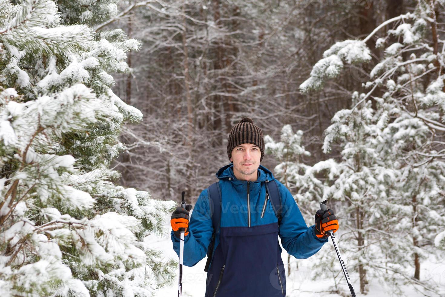 Skier with a backpack and hat with pompom with ski poles in his hands on background of a snowy forest. Cross-country skiing in winter forest, outdoor sports, healthy lifestyle, winter sports tourism. photo
