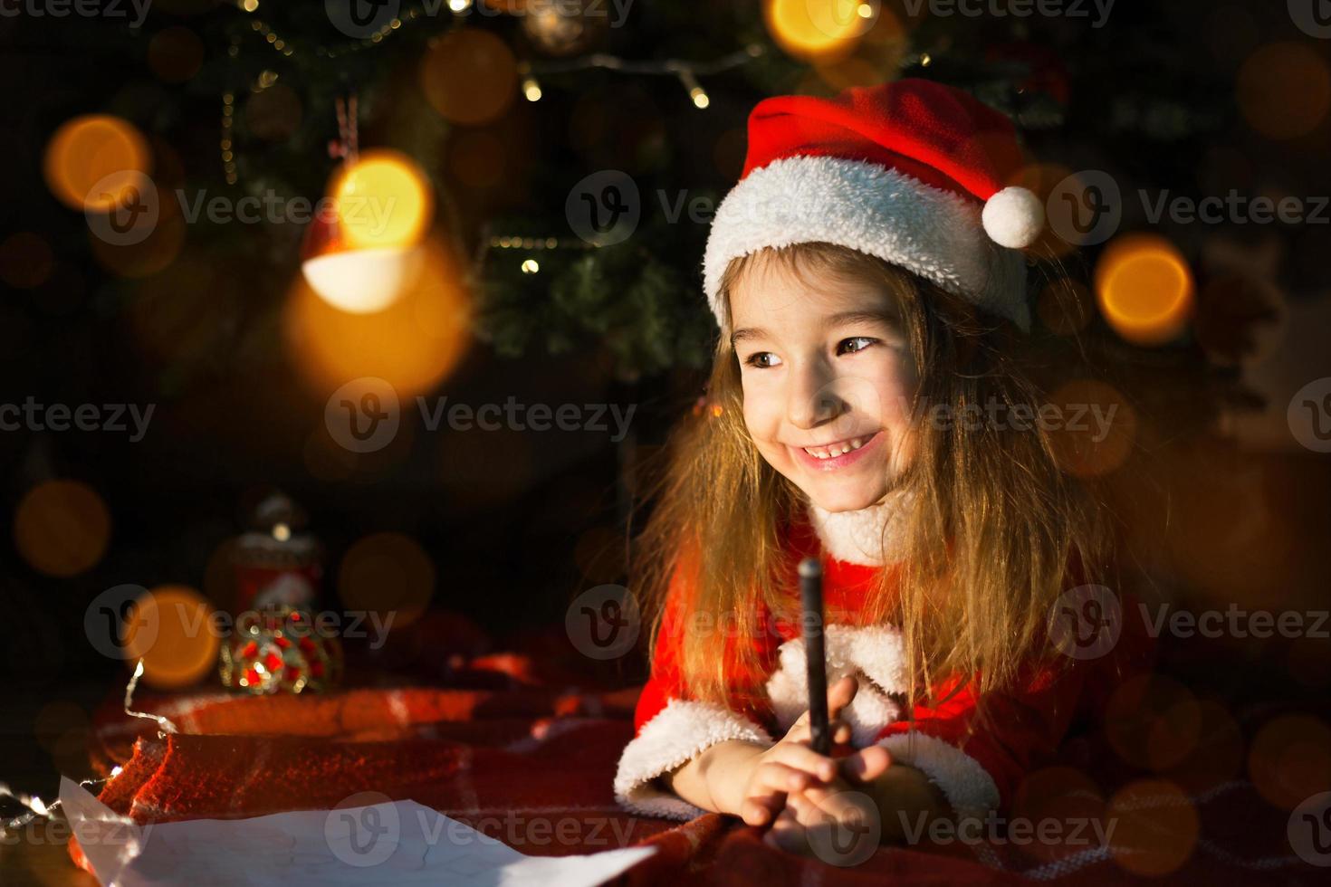 Little girl in a Santa hat and red dress under Christmas tree is dreaming, waiting for the holiday, lying on a plaid blanket. A letter on piece of paper, gifts. New year, Christmas. defocus lights photo